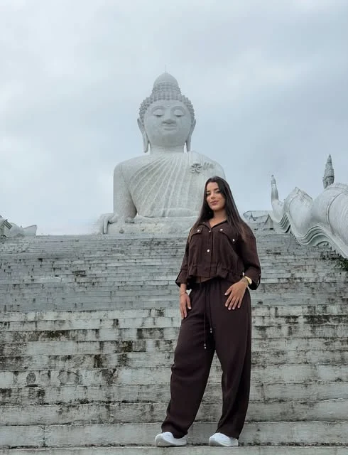 Bia Contrera de conjunto marrom na escadaria de um templo, com uma estátua de Buda ao fundo